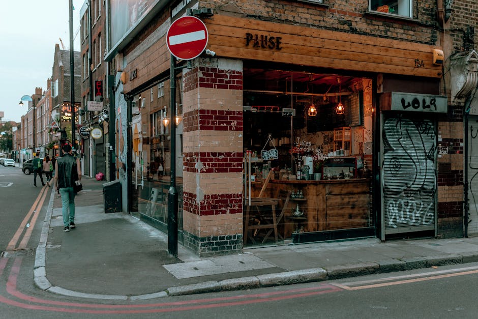 A busy urban street scene in Shoreditch, featuring multi-storey brick buildings with large windows and an extensive mural of a heron painted on the side of a building. The mural showcases a detailed, realistic depiction of a heron with textured feathers in shades of grey and white, set against a weathered brick wall. Below the mural, there is a graffiti-covered section with various tags and designs, some in bold black and white, others with colorful accents. The street is populated with pedestrians walking along the pavement, some engaged in conversation or looking at their phones. On the right side, a row of bicycles is parked along the curb, with a few secured to metal stands. The lighting appears overcast, casting diffuse light over the scene. Waste collection or rubbish removal services are not directly visible but are implied by the typical city street environment. This scene emphasizes the urban character and street art culture of Shoreditch, aligning with the context of alternative waste handling and private rubbish collection in the area, as managed by Waste Collection Shoreditch.