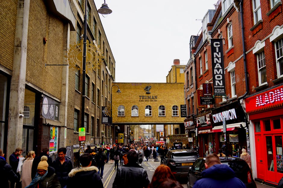 A busy urban street scene in Shoreditch showing a densely populated pedestrian area with many people walking along the pavement. On the left, there are multi-storey buildings with a variety of textures, including brick and concrete facades, some with large windows and iron street lamps hanging overhead. To the right, storefronts feature brightly lit signs for businesses such as a restaurant named 'Cinnamon' and other retail outlets, with illuminated signage and colorful branding. In the background, a yellow brick building with arched windows and the word 'TRUMAN' painted on it spans across the street, creating a distinctive skyline element. Vehicles, including cars, are parked or moving along the road, which is lined with street furniture such as signage, bollards, and parking meters. The overcast sky casts diffuse natural light over the scene, typical of a bustling commercial district. The image, associated with waste collection and possible local disposal services, captures the lively atmosphere of a popular, high-traffic area with various commercial and pedestrian elements visible, reflecting an environment where independent waste handling may be necessary for traders and shop owners.