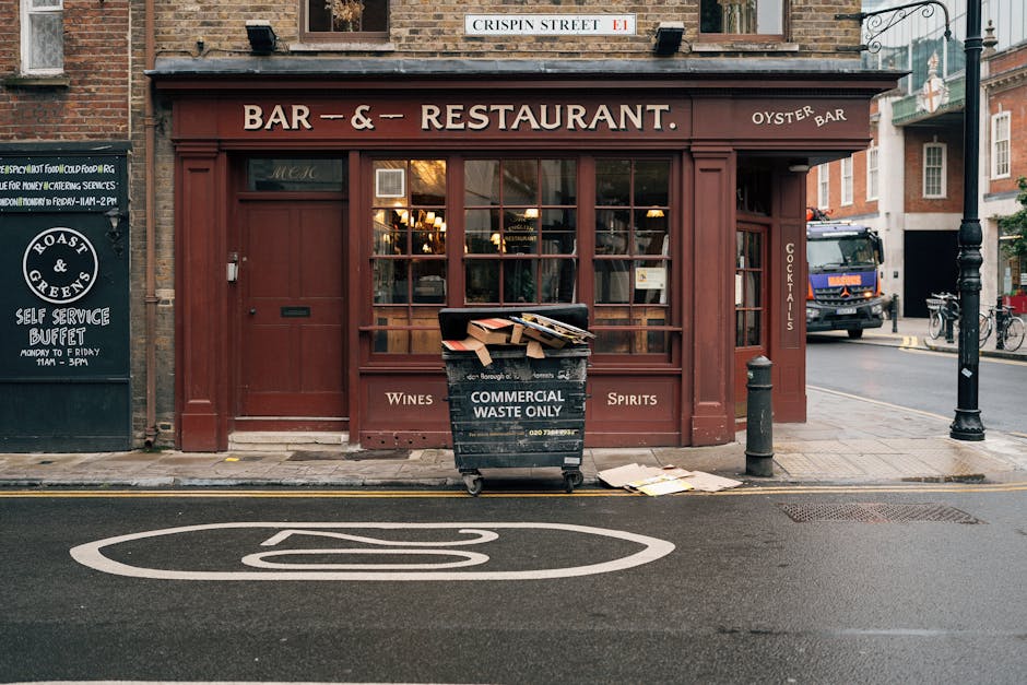 A busy urban street scene in Shoreditch showing a densely populated pedestrian area with many people walking along the pavement. On the left, there are multi-storey buildings with a variety of textures, including brick and concrete facades, some with large windows and iron street lamps hanging overhead. To the right, storefronts feature brightly lit signs for businesses such as a restaurant named 'Cinnamon' and other retail outlets, with illuminated signage and colorful branding. In the background, a yellow brick building with arched windows and the word 'TRUMAN' painted on it spans across the street, creating a distinctive skyline element. Vehicles, including cars, are parked or moving along the road, which is lined with street furniture such as signage, bollards, and parking meters. The overcast sky casts diffuse natural light over the scene, typical of a bustling commercial district. The image, associated with waste collection and possible local disposal services, captures the lively atmosphere of a popular, high-traffic area with various commercial and pedestrian elements visible, reflecting an environment where independent waste handling may be necessary for traders and shop owners.