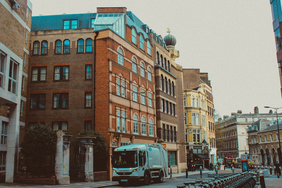 A street scene in an urban area featuring a row of multi-story buildings with a mix of architectural styles, including red brick facades, large windows, decorative cornices, and a dome-topped tower. In the foreground, a waste collection truck with a metallic blue finish is parked on the pavement near the curb, surrounded by a small enclosed area with stone pillars and an iron gate. The street is lined with bollards and bicycle racks, with some bicycles secured to the racks. Overcast lighting creates a soft, neutral atmosphere, and the background shows additional historic buildings with classical detailing, conveying a typical cityscape that may be serviced by private waste collection firms such as Waste Collection Shoreditch for rubbish removal or site clearance, reflecting an environment where independent waste disposal methods are in use outside local authority services.